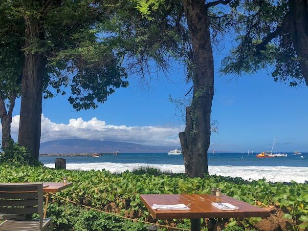 Aloha Mixed Plate View - Maui. Two tables are shown overlooking the beach and ocean with boats floating in the water. 