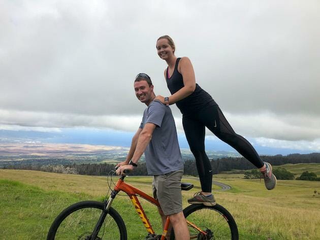 Man and Woman posing on bike with the woman standing on the back of the bike. View in the background of Maui. 