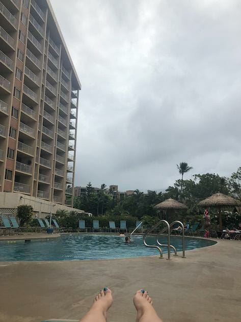 Royal Kahana Maui Pool with feet in the photo. Toenails are painted blue. Hotel building is in the background. Sky is gray and cloudy. 
