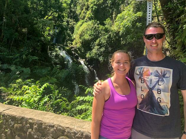 Man and Woman posing for photo in front of stone wall with small waterfall and thick green foliage in the background. 