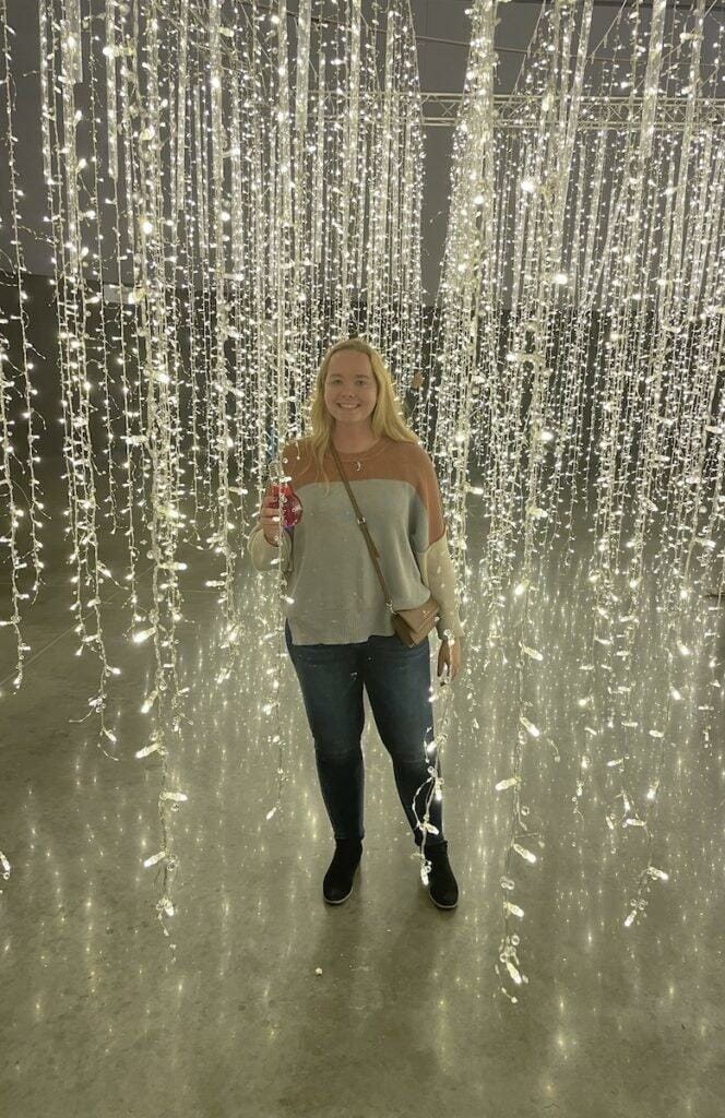 Young woman wearing an orange and grey sweater, jeans, and black booties. Holding a red drink in her hand. Around her, Holiday Lights are hanging down from the ceiling.