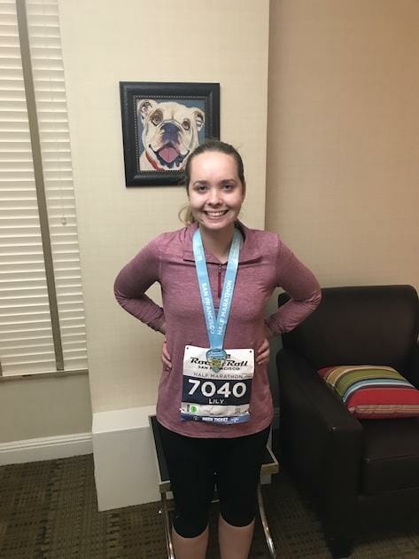 Goals for 2022 - Run Half Marathon: A young woman is posing with a half marathon medal and a running bib. A painting of a dog is hanging behind her. 