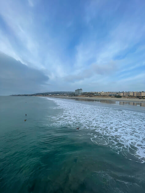 View of Pacific Beach (PB) from the Pacific Beach Pier. Cloudy sky and surfers on the water. Lots of buildings in the distance. 