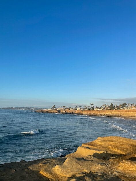 View of San Diego from sunset cliffs in Point Loma. Waves are crashing and palm trees, homes and mountains can be seen in the distance. 