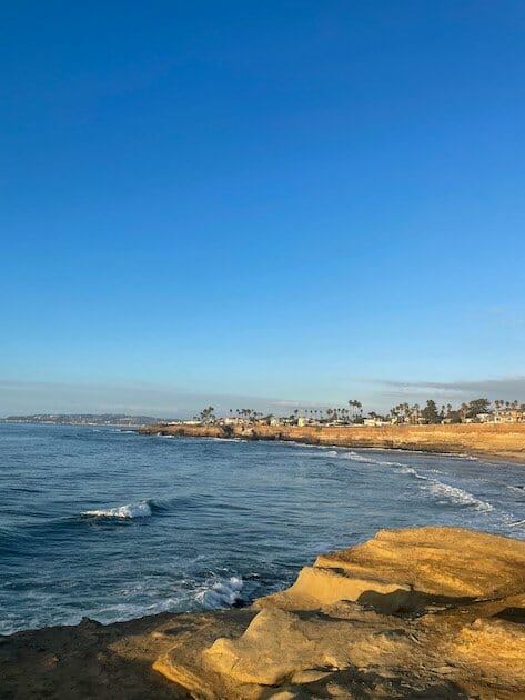 View of San Diego from sunset cliffs in Point Loma. Waves are crashing and palm trees, homes and mountains can be seen in the distance.