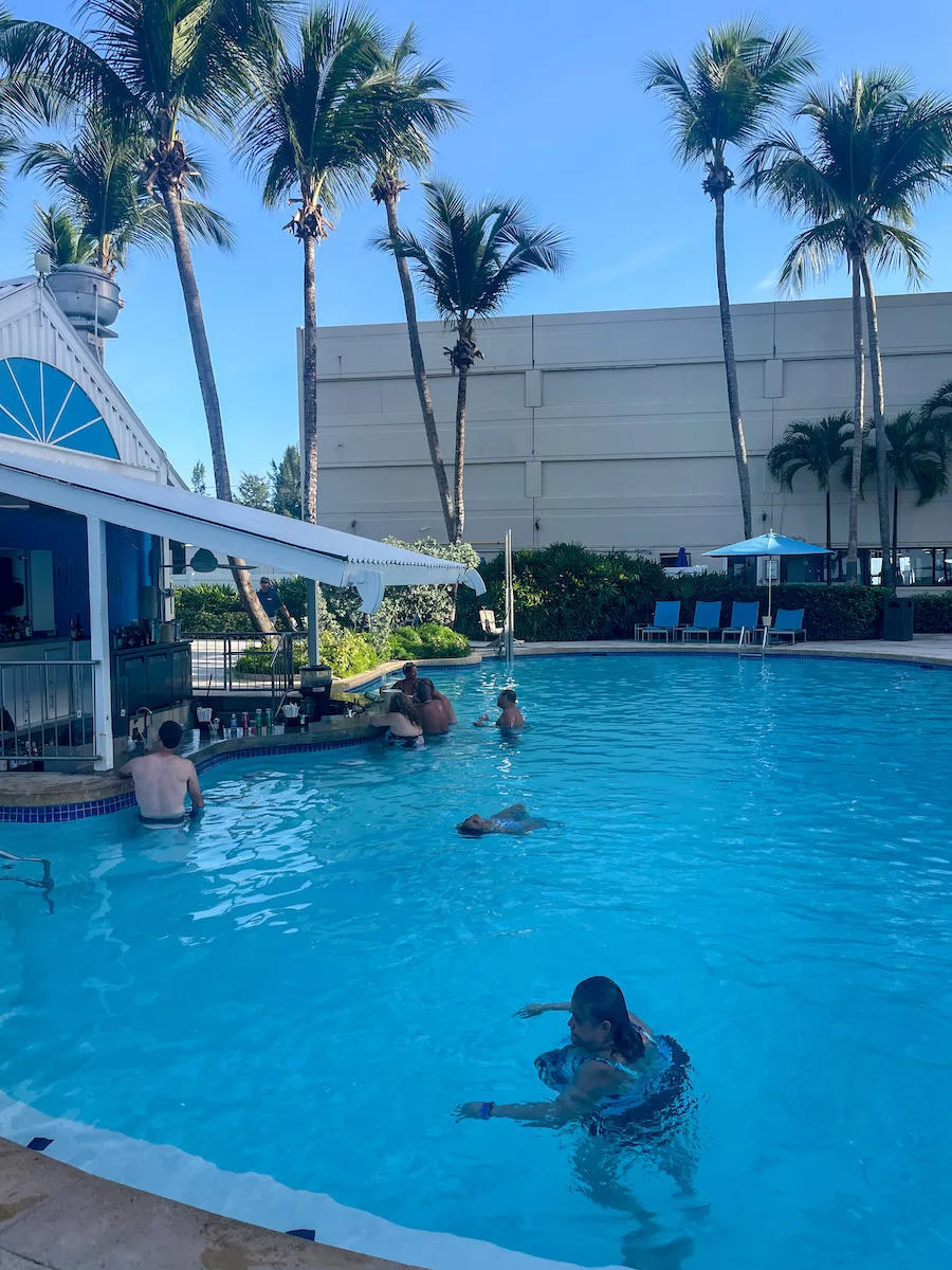 Bar in pool with a few people, surrounded by palm trees. 