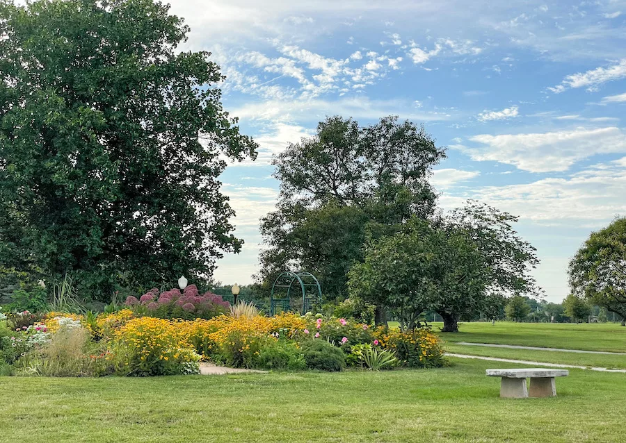 View of a colorful flower garden surrounded by grass and trees, with a cloudy blue sky. 