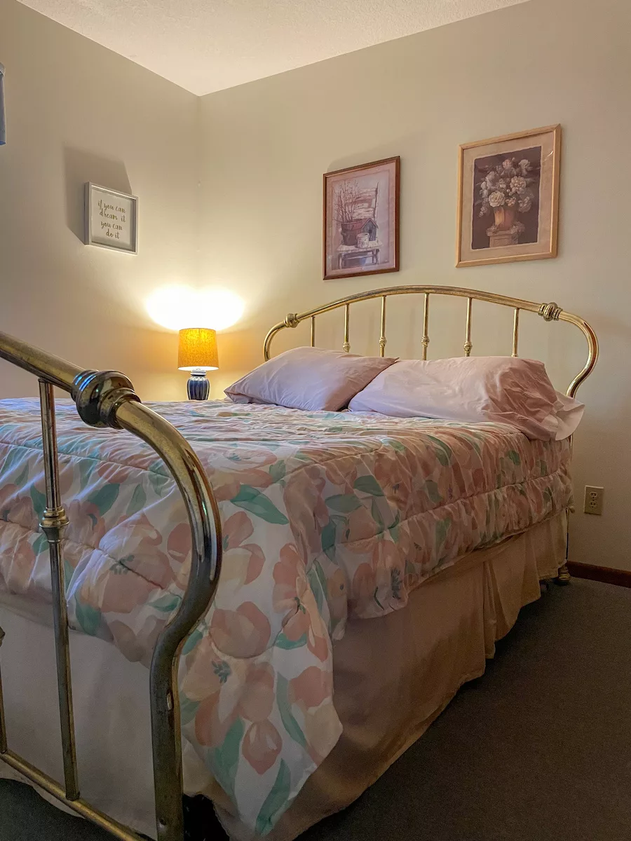 Bedroom of a cottage with a made bed and photos hanging on the wall. There is a lamp turned on in the corner. 