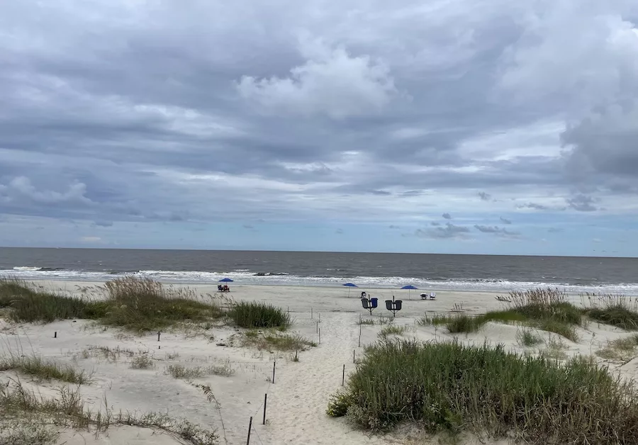 Image of half sand dunes, half clouds and light blue sky, with a little bit of ocean sandwiched inbetween the two. 