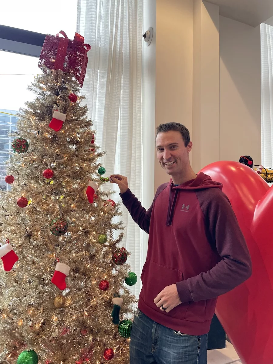 Man standing next to white Christmas Tree holding a pickle ornament and smiling