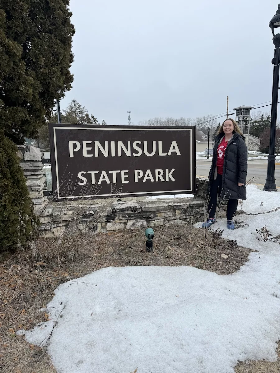 Peninsula State Park Entrance sign with you woman posing next to it in the snow. 
