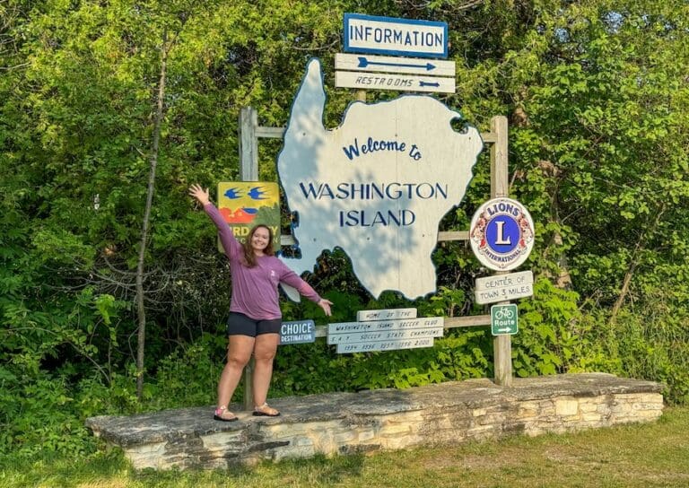 Image of young woman in long-sleeved purple t-shirt and shorts with Welcome to Washington Island sign in Door County, Wisconsin