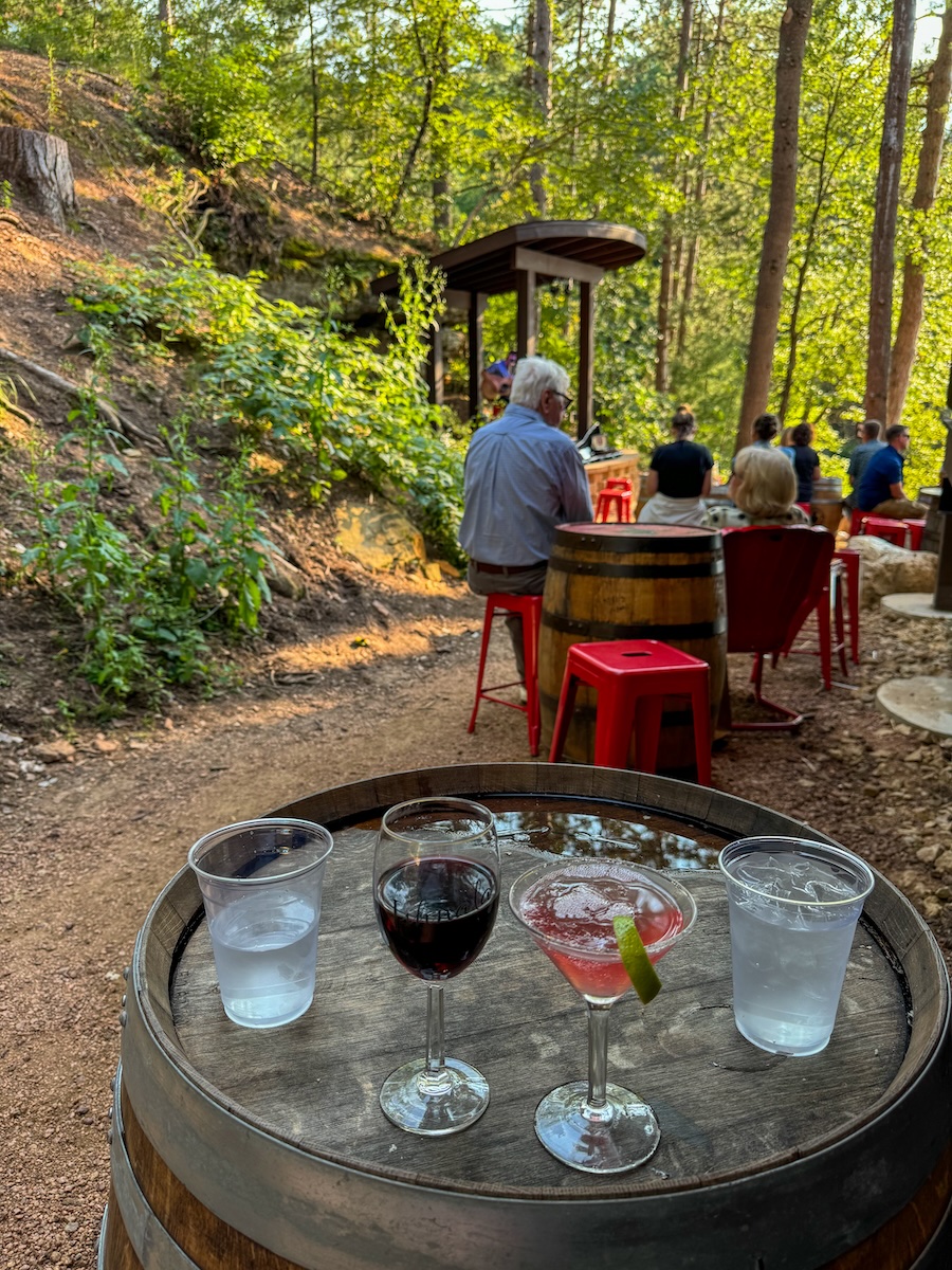 Image of a barrel outside with a class of wine, a cocktail, and two glasses of water on top of it. The barrel is situated in the woods surrounded by green trees.