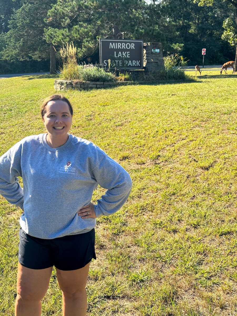 Image of a young woman wearing hiking shorts and a gray sweatshirt standing in front of a sign to Mirror Lake State Park - Wisconsin Dells, WI
