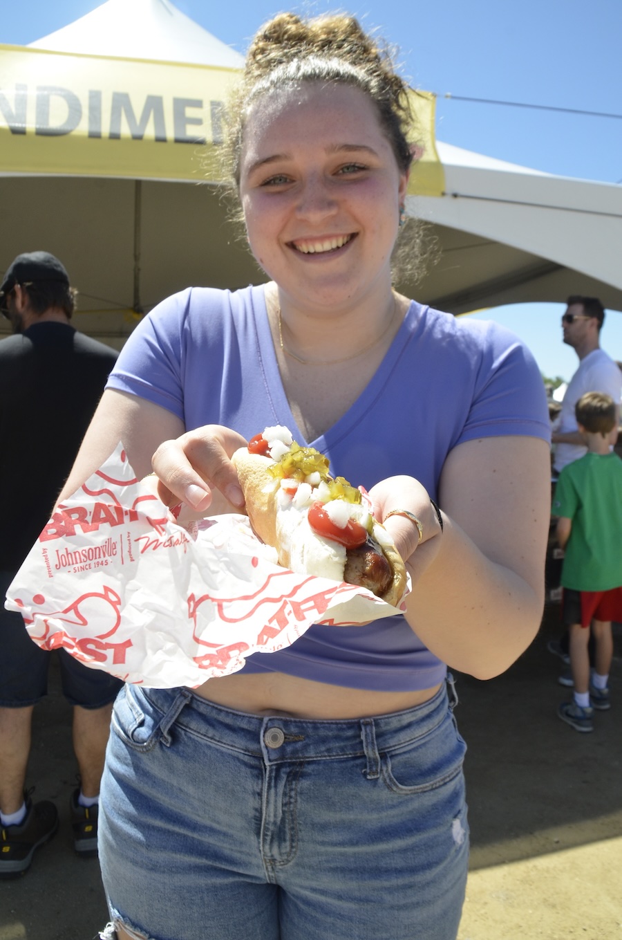 Image of young woman wearing a purple shirt and blue jeans eating a load brat in Madison, WI